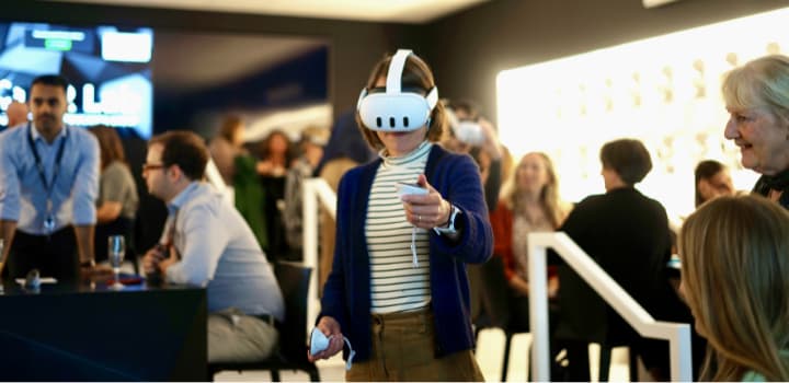 A woman wearing a white VR headset and holding controllers tries the Rural Minds experience at its launch event, with a small crowd around her.