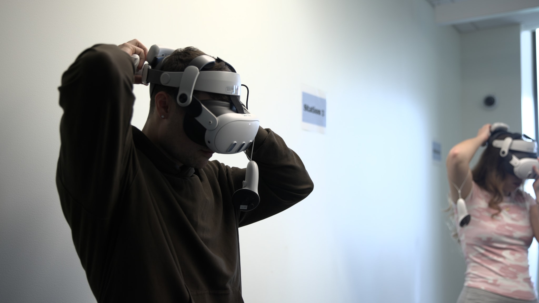 Two nursing students pull VR headsets over their heads in a classroom, standing beside a wall sign reading Station 3.