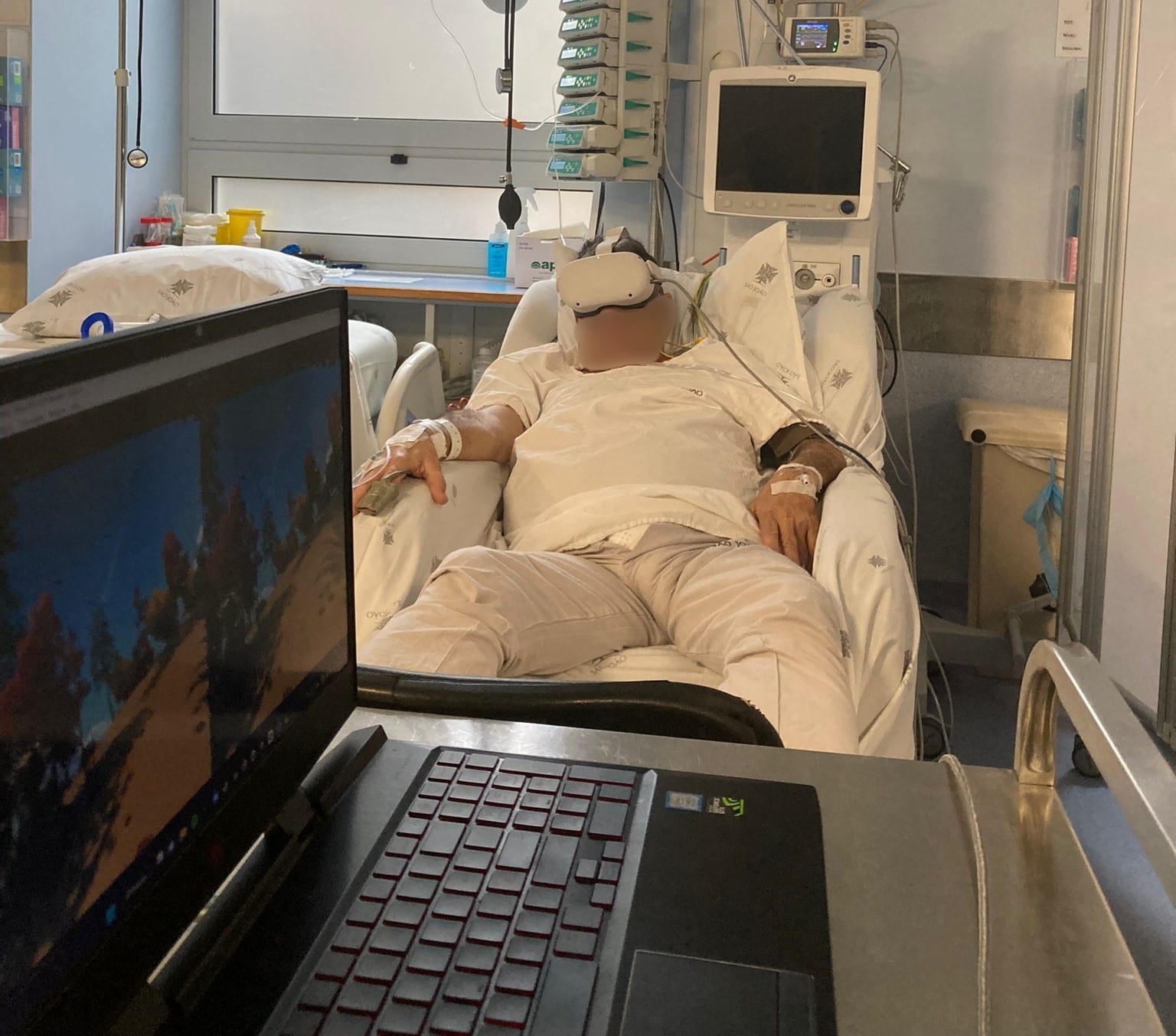 A patient reclines in a hospital bed wearing a white VR headset, with medical monitors and a laptop nearby.