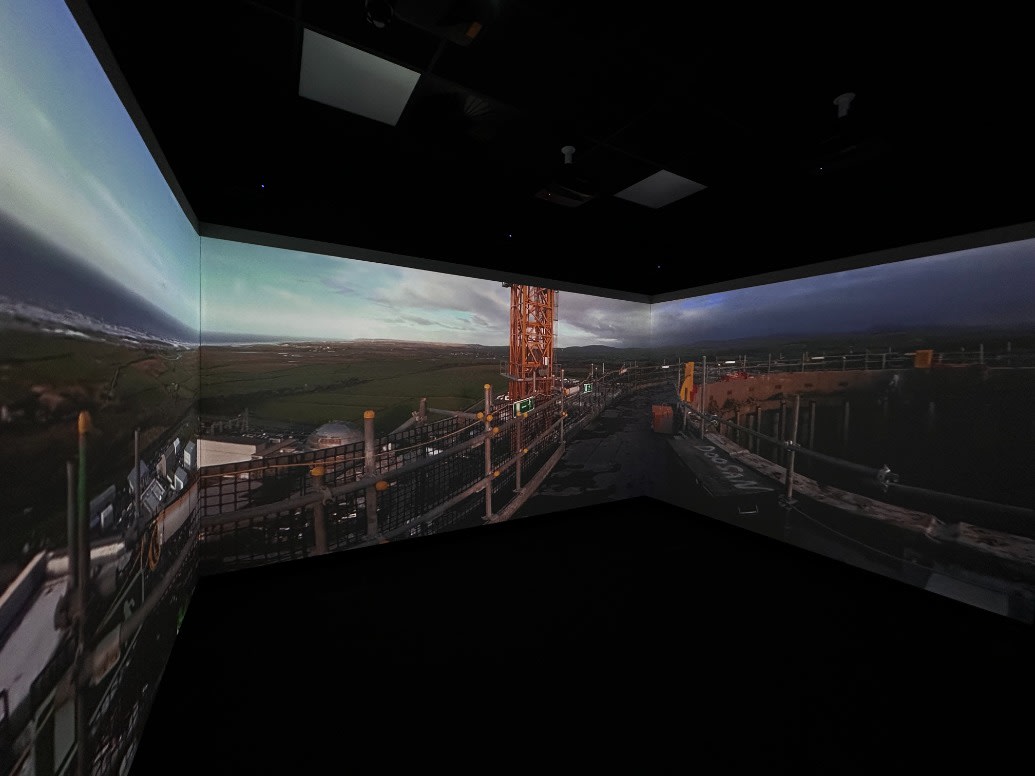 A darkened room with a wide 360-degree panorama of a Sellafield site projected across three walls, showing gantry walkways, an orange lattice tower and the Cumbrian coastline under a cloudy sky.