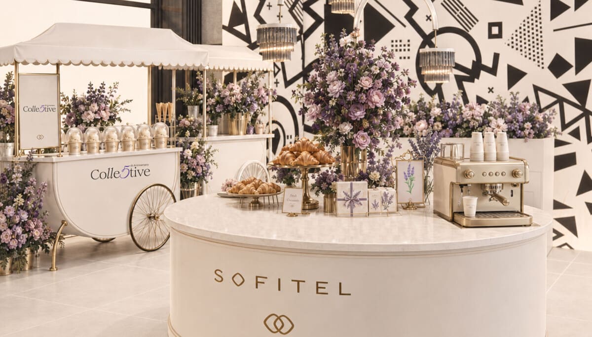 AI-generated rendering of a white Sofitel-branded counter with croissants, flowers and an espresso machine, beside a Collective fifth-anniversary cart against a black-and-white geometric wall.