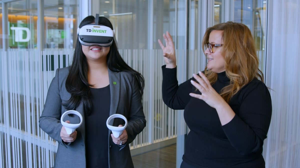 A TD colleague wearing a white VR headset branded TD Invent and holding two controllers, while another colleague gestures beside her inside a TD office