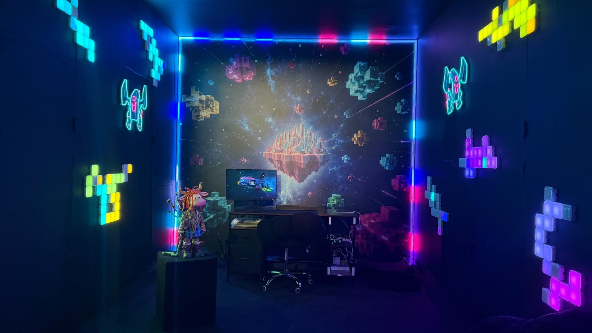 A dark installation room lit by colourful LED wall tiles and neon character outlines, with a cosmic backdrop, a desk with a monitor and chair, and a character figure on a plinth.
