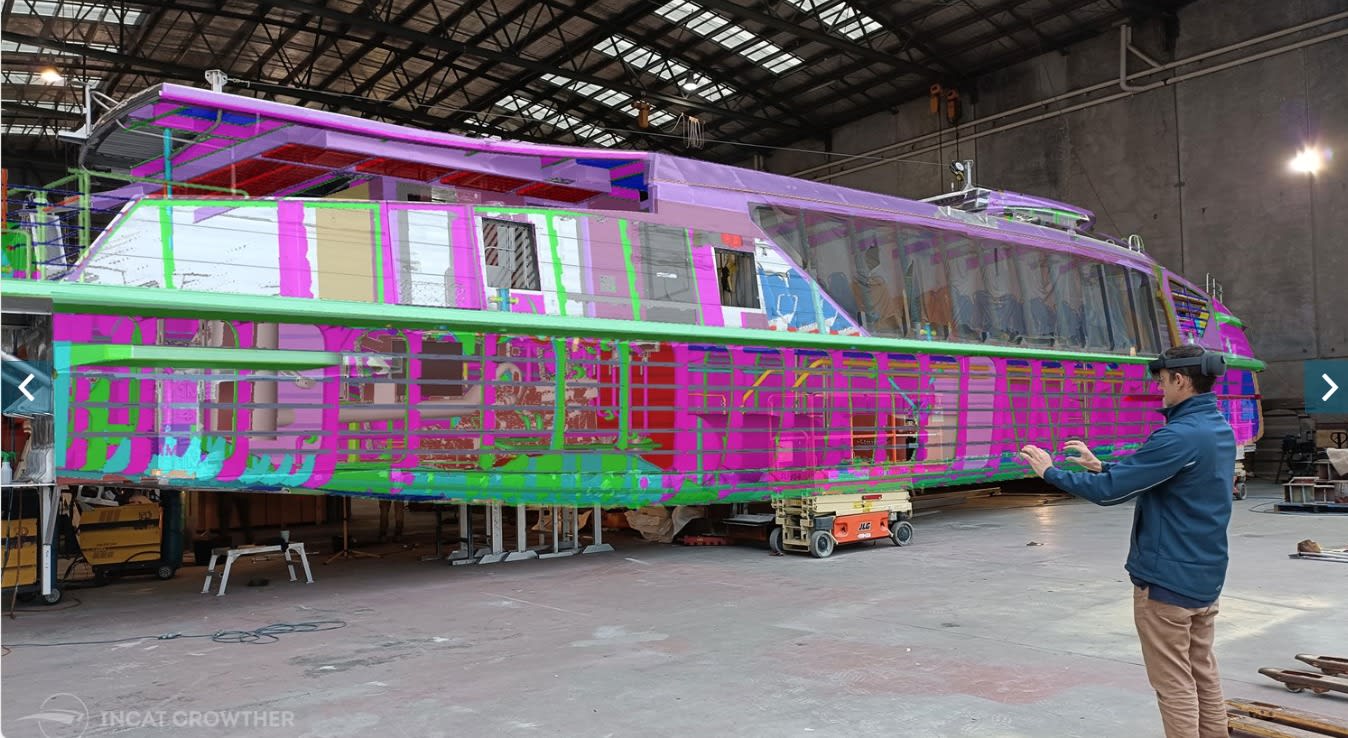 Engineer wearing a mixed-reality headset on the shipyard floor beside a part-built ferry, with the coloured 3D structural model overlaid on the physical hull