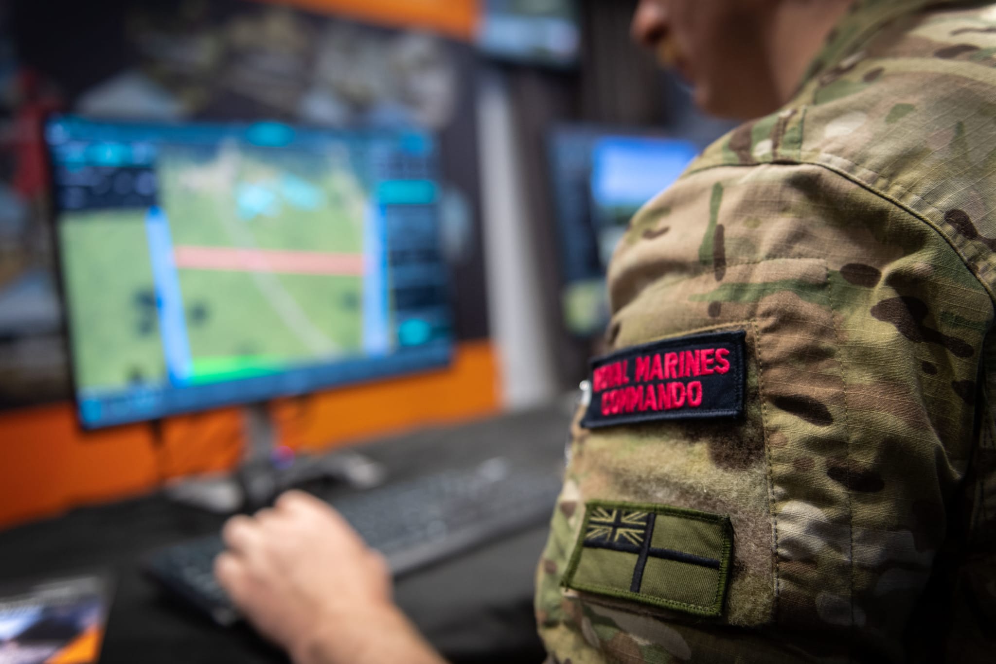 A Royal Marines Commando in camouflage uniform operating the DVS2 simulation at a desktop workstation, with a tactical map on the monitor in front of him