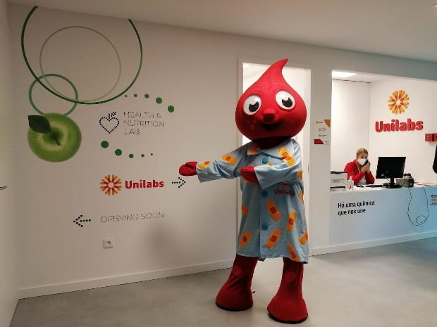 A red blood-drop mascot stands beside the Health & Nutrition Lab wall and a staffed Unilabs reception desk.