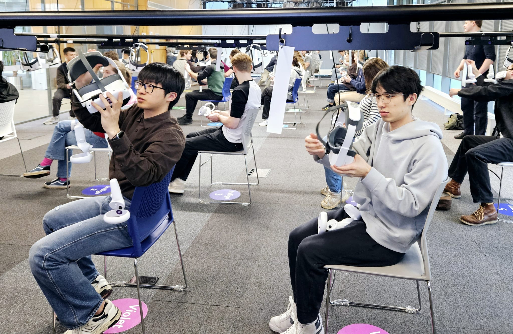 Students seated in rows in a bright classroom lift white VR headsets that hang on cables from overhead ceiling rails, controllers in their laps and coloured position markers on the floor.