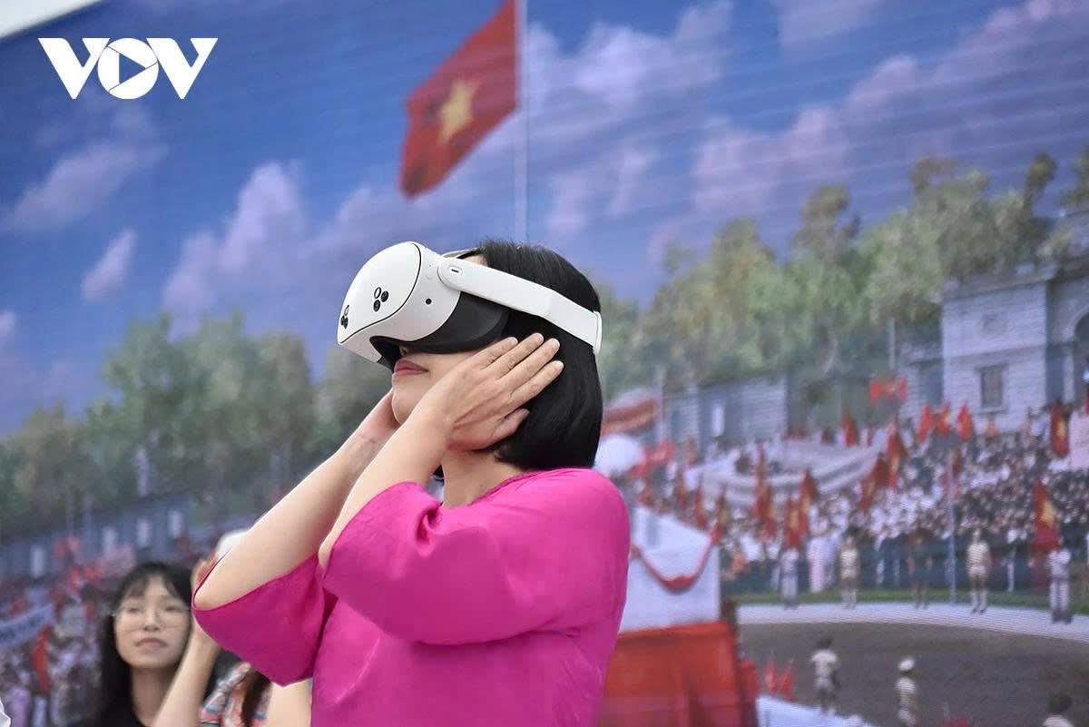 A visitor wearing a white Meta Quest headset raises both hands to the device, standing in front of a large mural of crowds and flags at Ba Dinh Square.