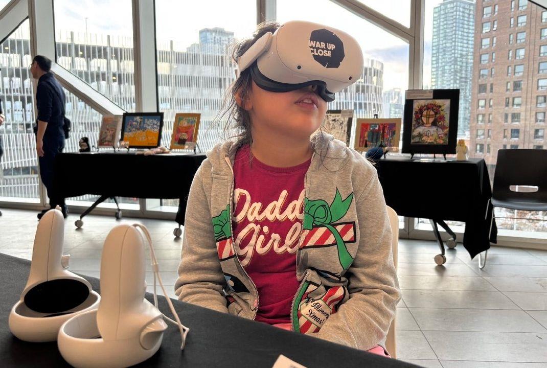 A child seated at a table wears a white VR headset branded 'War Up Close', with two white hand controllers on the table in front