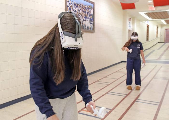 A student in a white VRNA-branded VR headset holds a ring controller in a school hallway while another headset-wearing student stands behind her.