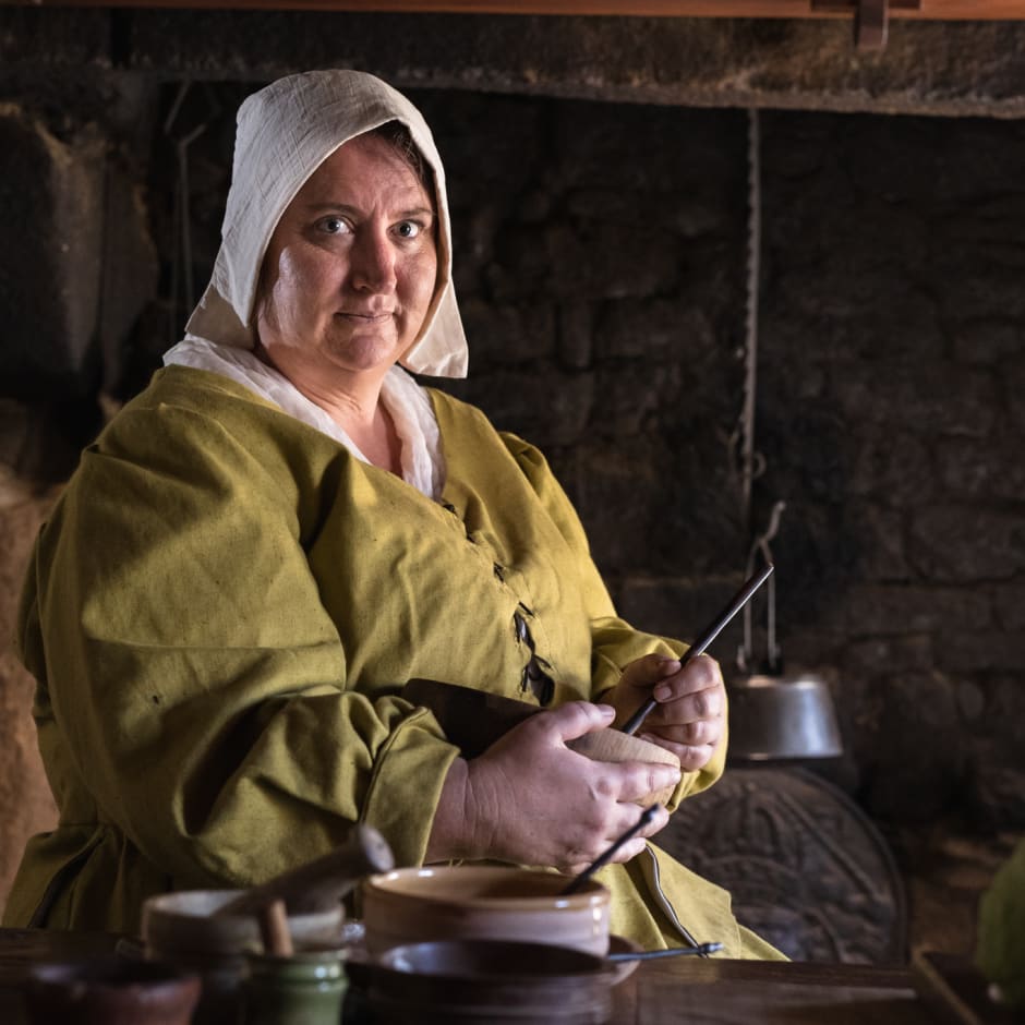 Women sits on stool next to table, she is dressed as a housekeeper from 1600s