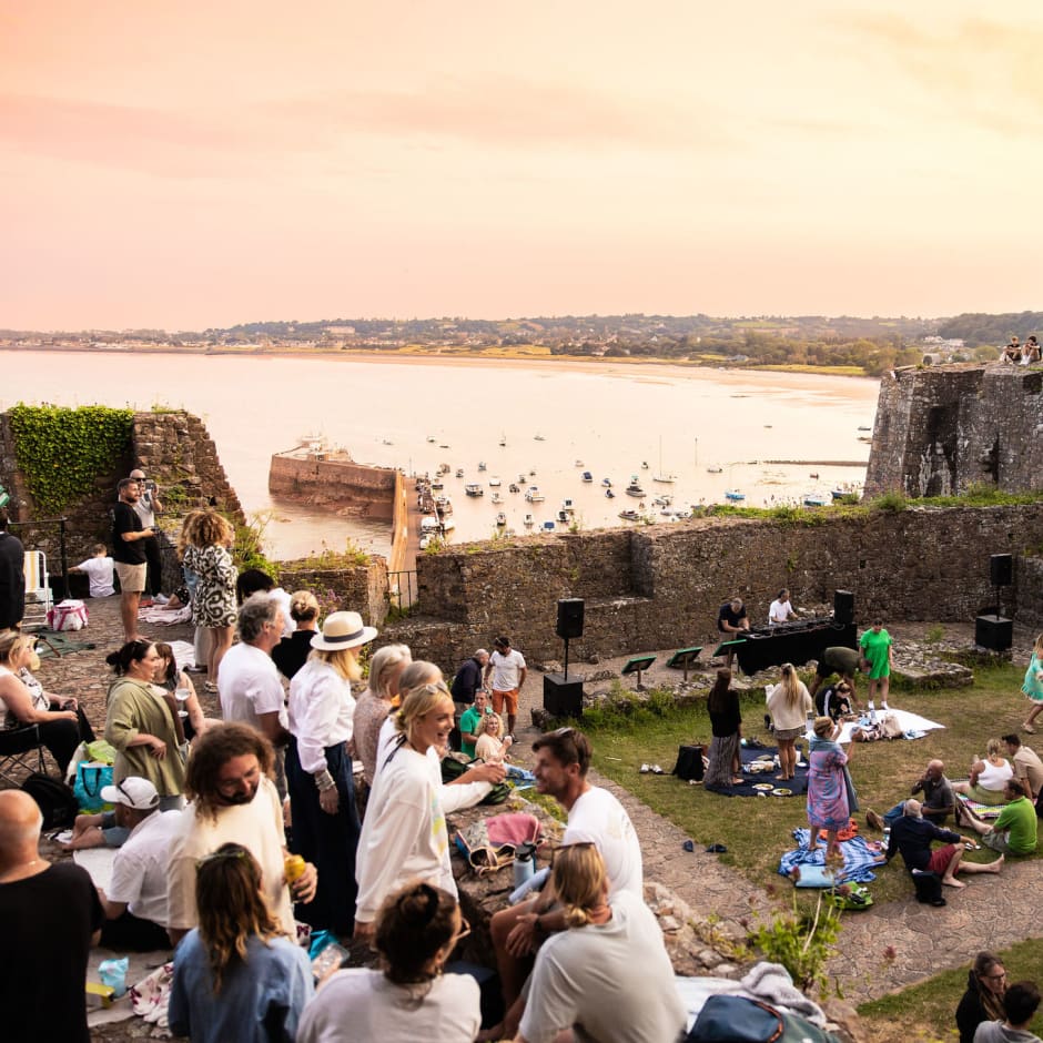 A lot of people fill an outdoor area with the sea in the background at sunset