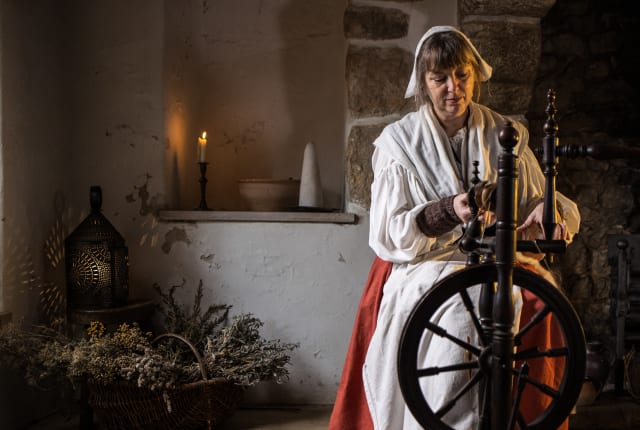 Women in 17th century costume sits at a spinning wheel