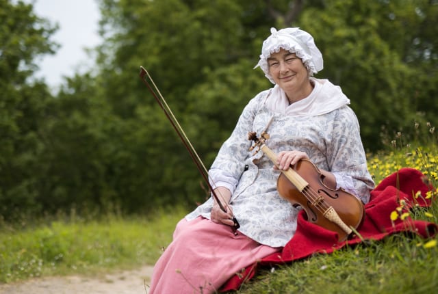 La Hougue Bie Mary Le Brun woman in period costume sits on grass holding a violin