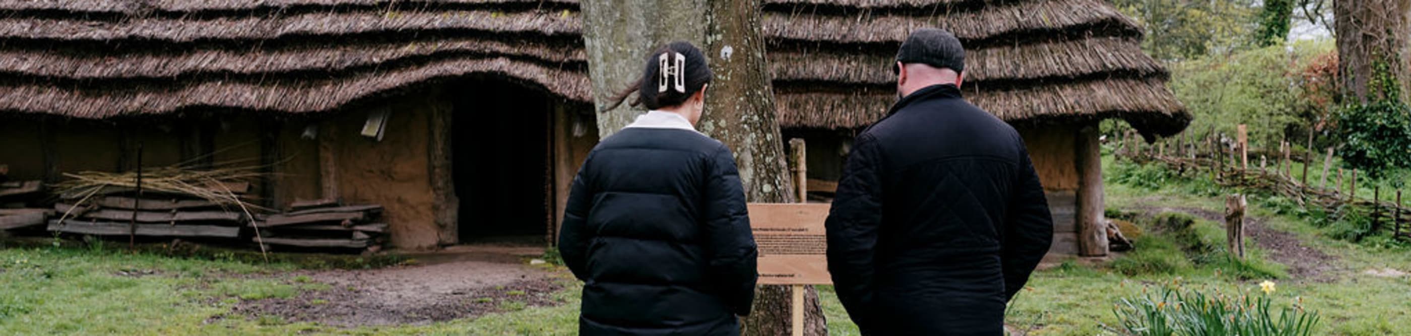 Two people read sign inf front of a replica neolithic longhouse