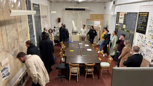 A large group of people stand around a long table with maps on the wall