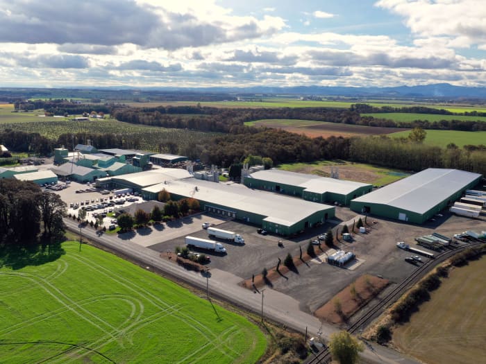 Aerial view of a Smith Seed facilities surrounded by farmland and trees, featuring multiple green-roofed buildings, parking lots with vehicles, semi-trucks, and a backdrop of distant mountains beneath a partly cloudy sky.