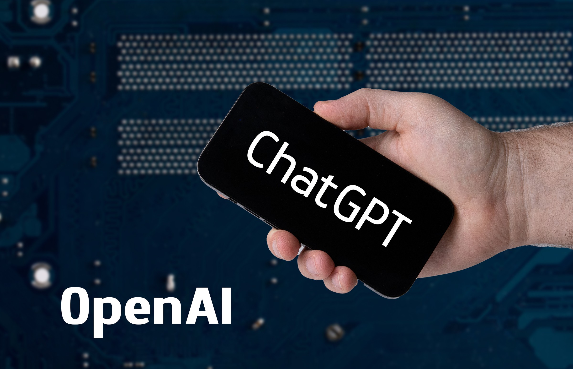 A hand holding a black smartphone sideways with the white ChatGPT wordmark on screen, in front of a dark blue circuit board, with the word OpenAI in white in the lower left