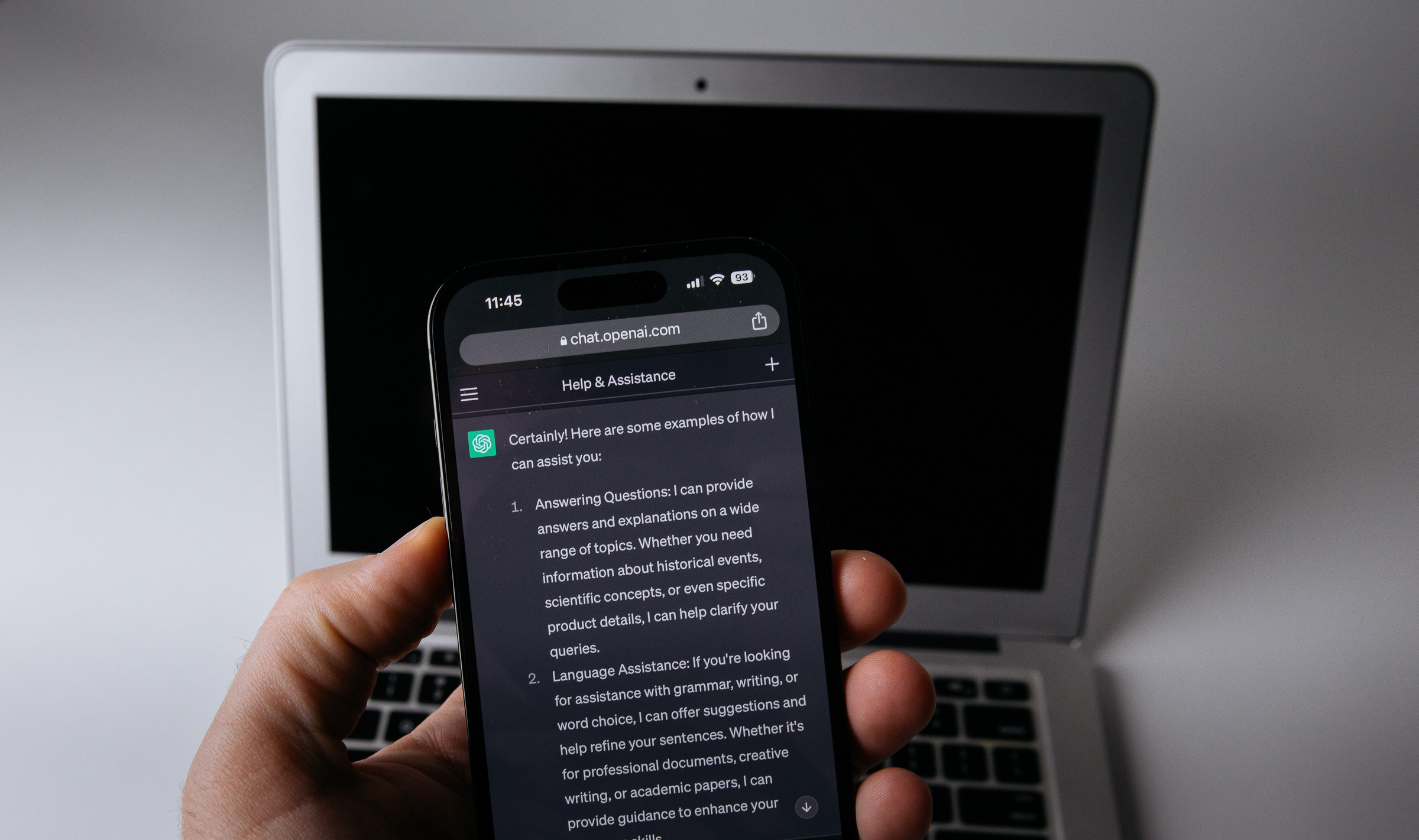 A hand holding a smartphone showing a ChatGPT reply on chat.openai.com that begins Certainly! Here are some examples of how I can assist you, with a silver laptop behind it
