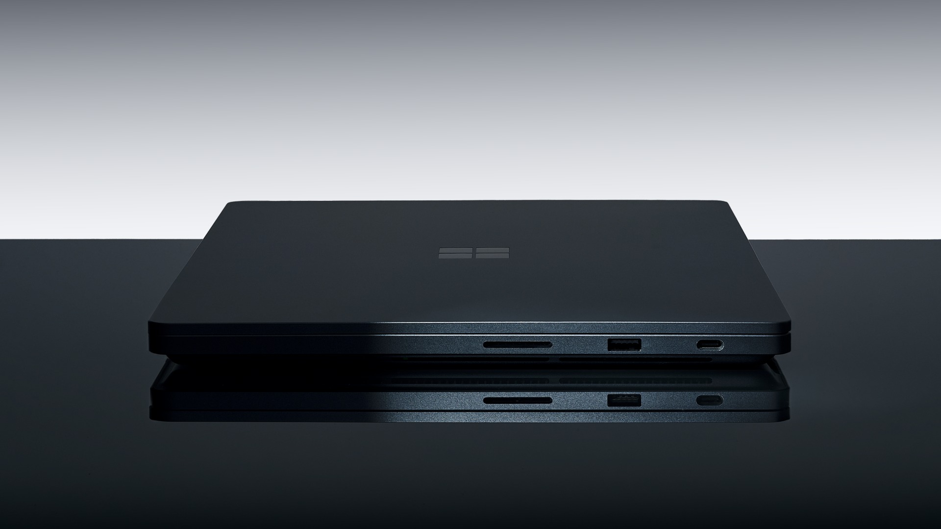 Surface Laptop Ultra closed on a dark reflective table, side view showing the Windows logo on the lid, a full-size SD card slot, a USB-A port and a USB-C port