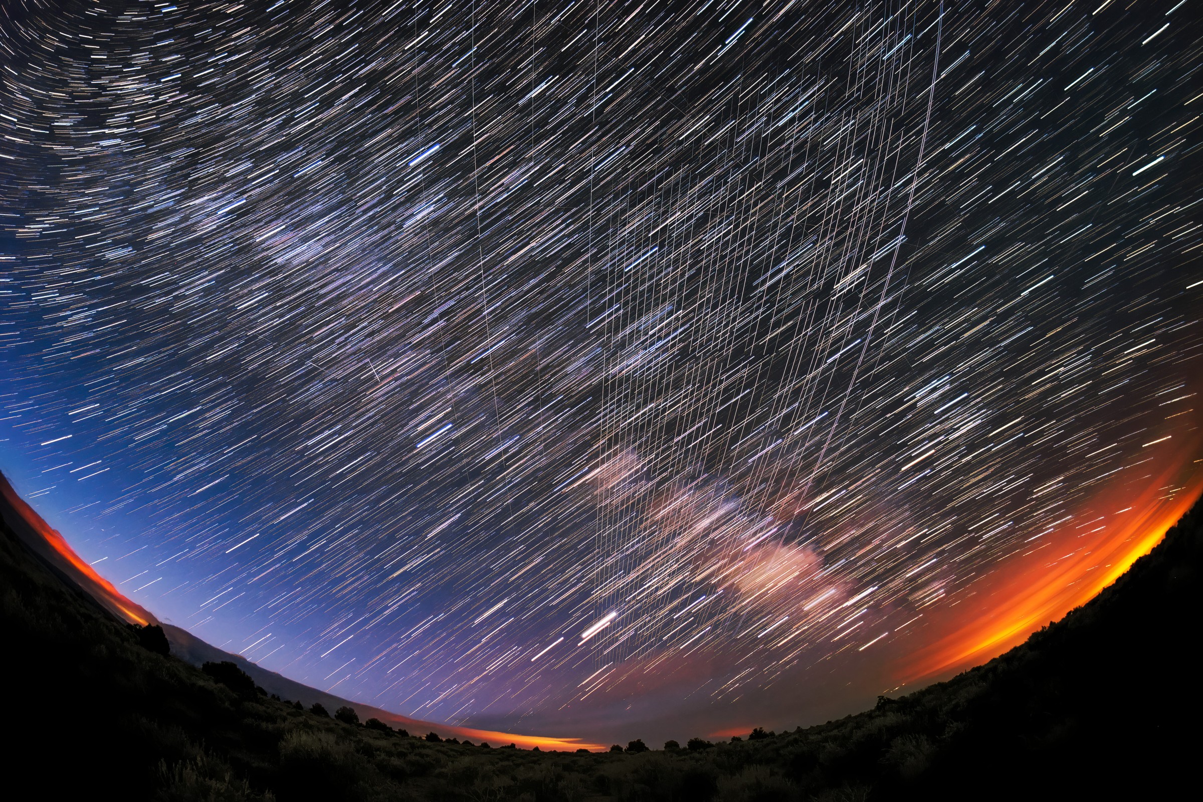 Long-exposure night photo over New Mexico in which a bundle of straight white Starlink satellite streaks cuts across curved star trails above a dark hillside and an orange glow on the horizon