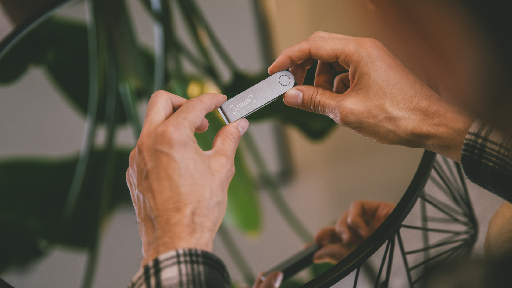 Official Ledger photo of two hands holding a silver Ledger Nano X hardware wallet with the Ledger logo, above a round glass table with a green plant in the background