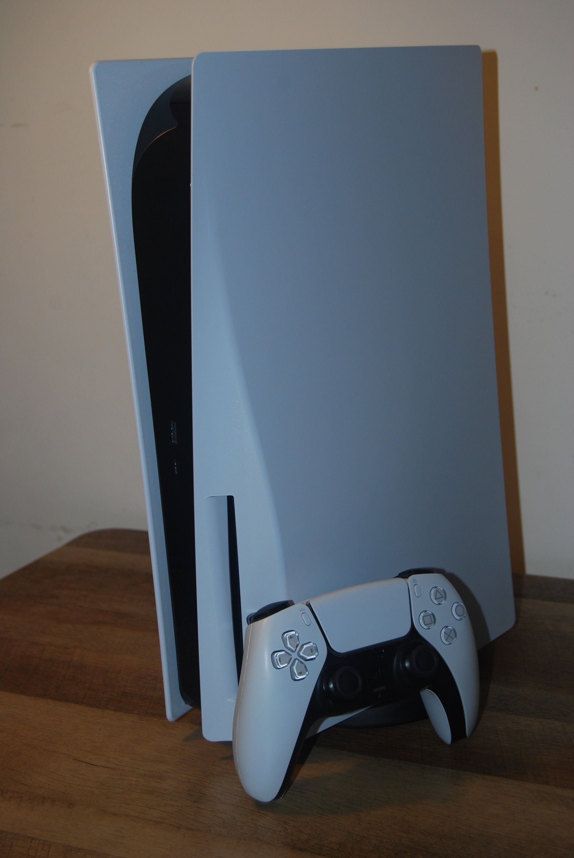 White standard PlayStation 5 console standing upright on a wooden table with a white DualSense controller leaning against it