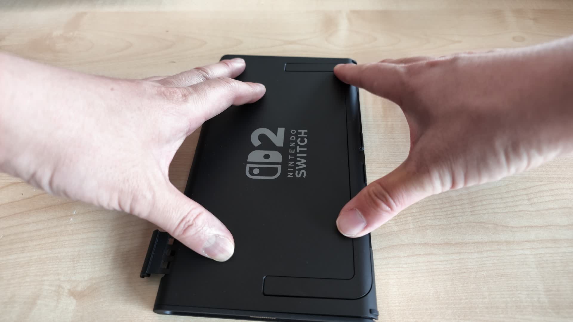 Two hands gripping the black back cover of a Nintendo Switch 2 console lying face down on a wooden table