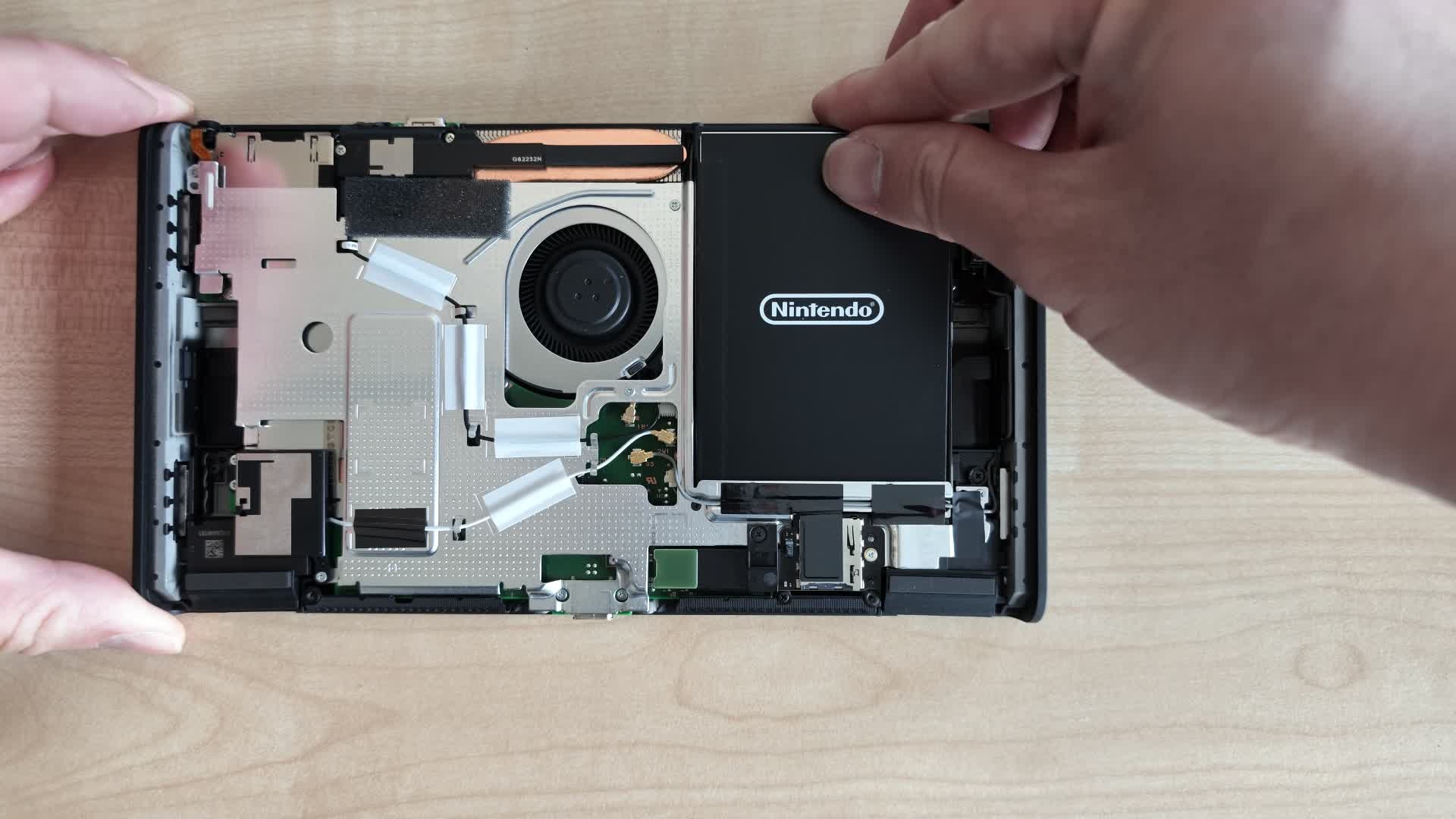 Hands lifting a black Nintendo-branded battery out of an opened Nintendo Switch 2 console, with the cooling fan and circuit board visible