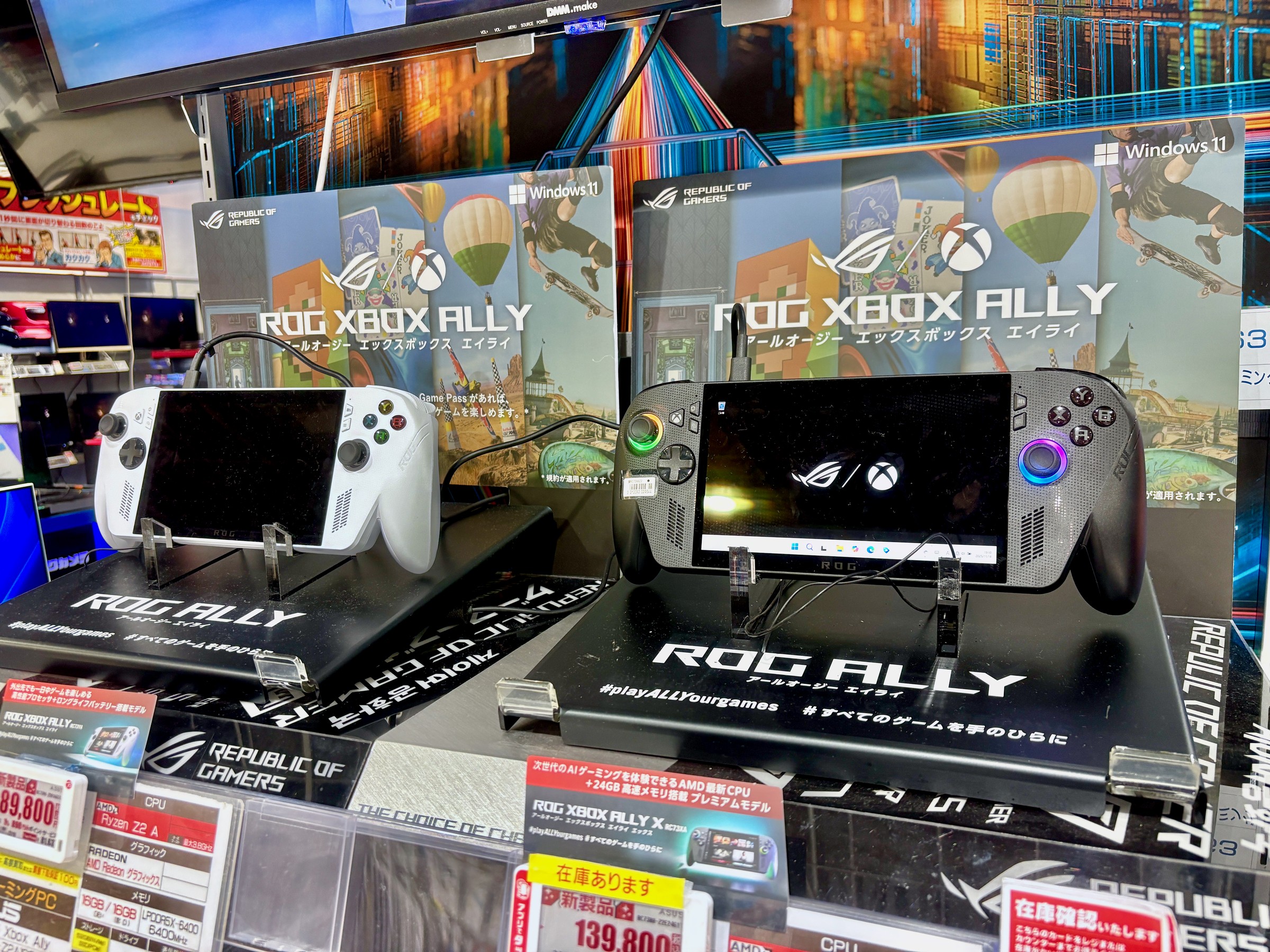 White ROG Xbox Ally and black ROG Xbox Ally X handhelds on display stands in a Japanese electronics store, with ROG Xbox Ally boxes behind them