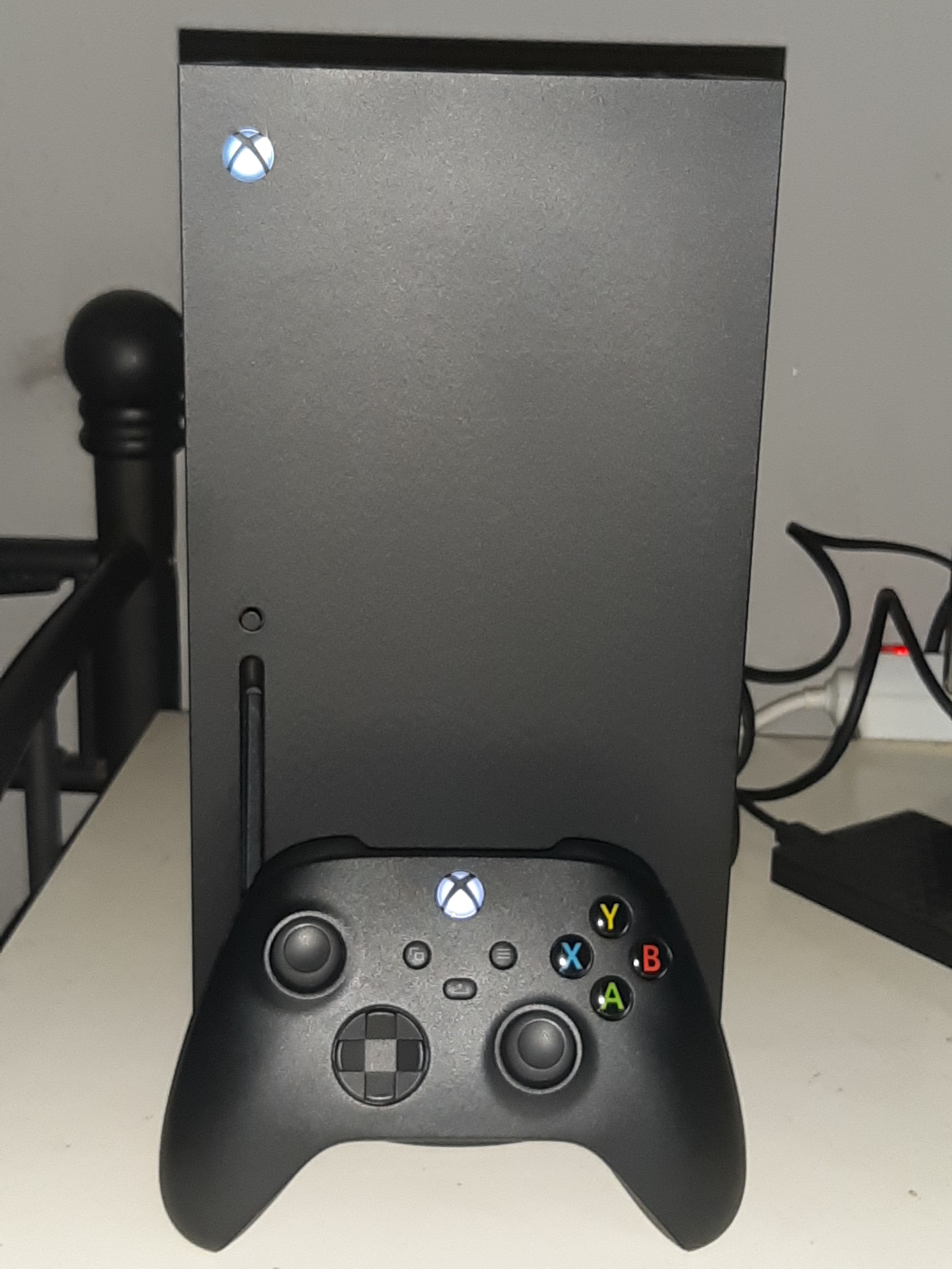 Black Xbox Series X console standing upright on a white shelf with a black Xbox Wireless Controller leaning against its front