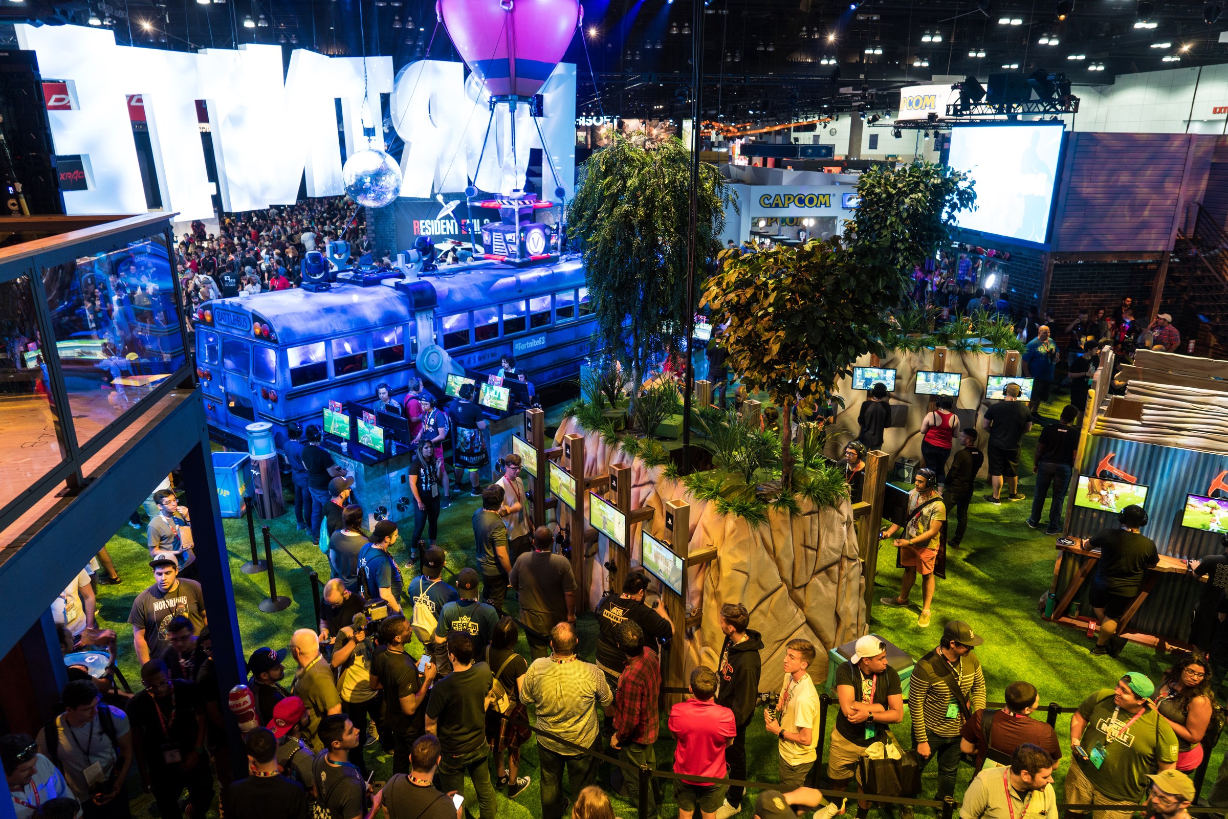 Crowds of players at Epic Games' Fortnite booth at E3 2018, with a giant purple balloon over a blue Battle Bus and rows of game stations set among trees and rocks