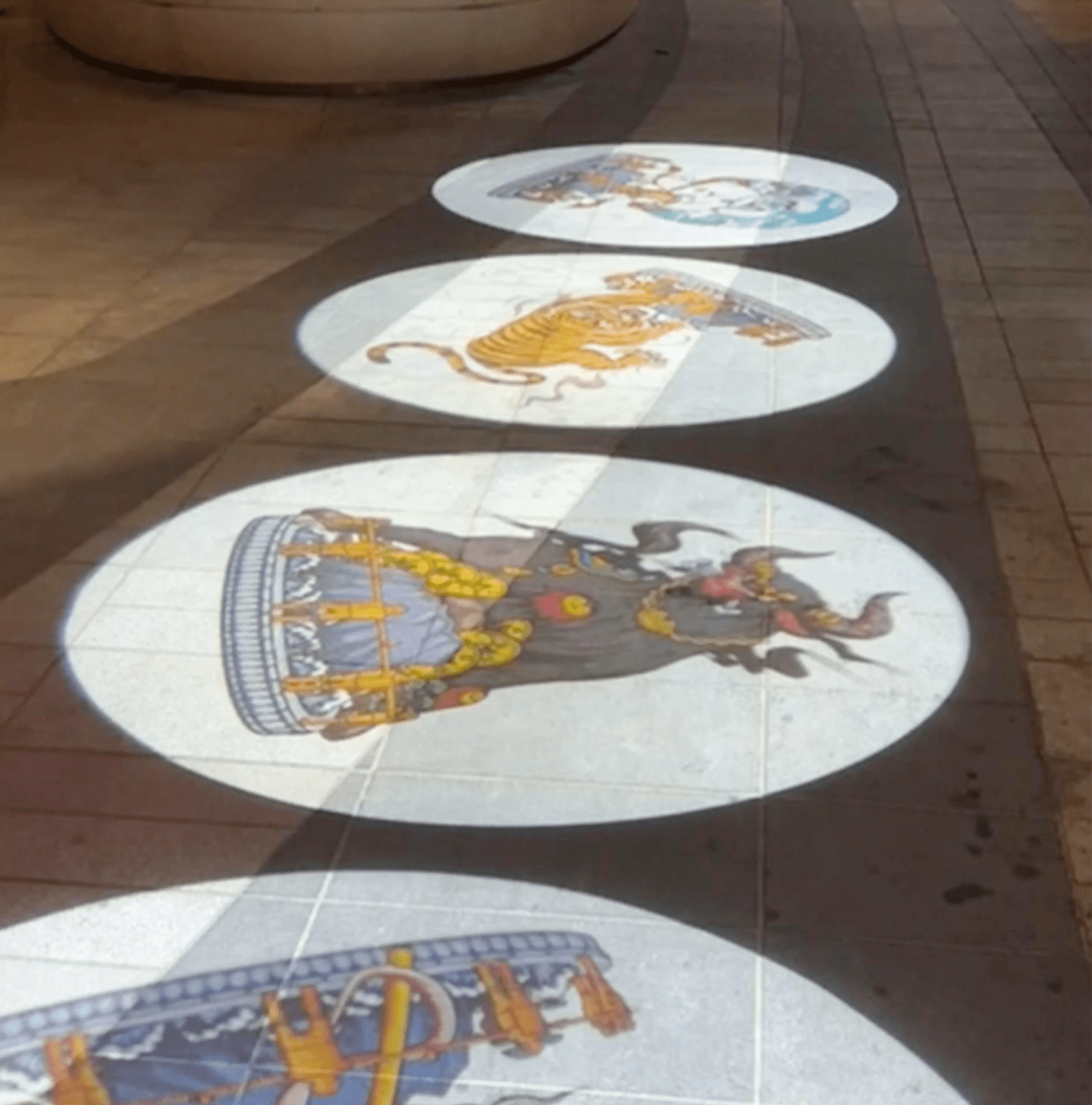 Traditional folk-art animals in bright circles cast by gobo lights along an outdoor paver walkway at night