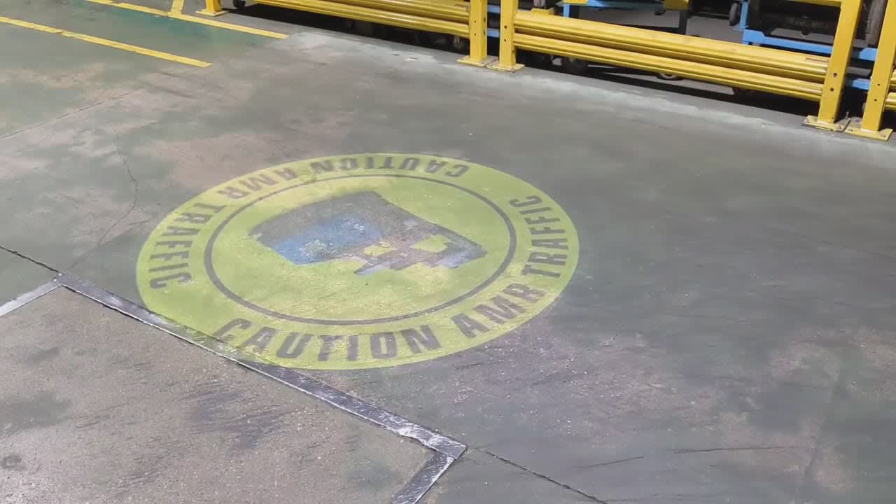 Rotating caution sign projected on a warehouse walkway next to guardrails — virtual safety signage projector