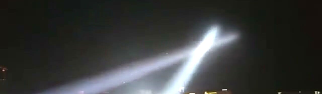 Two searchlight beams crossing in an X over a real night city skyline, cast from a rooftop install
