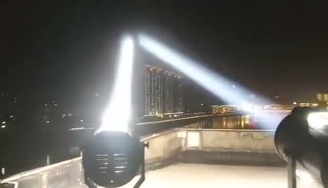 Two searchlight beams crossing over a real night city skyline from a rooftop install — Infinity Projectors outdoor sky searchlight