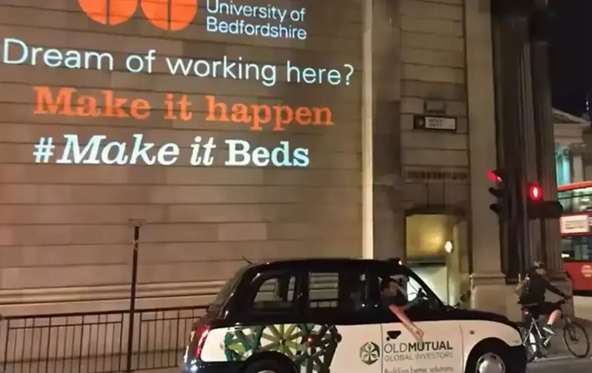 University recruitment message projected on a city-street building facade beside a passing bus — Infinity Projectors