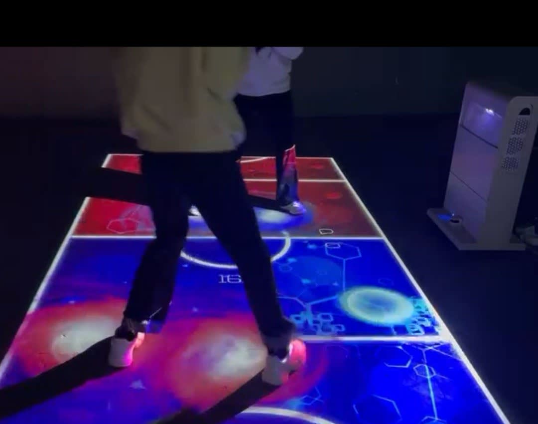 Video: two players step across a red and blue space game projected on a dark floor by a mobile interactive tower