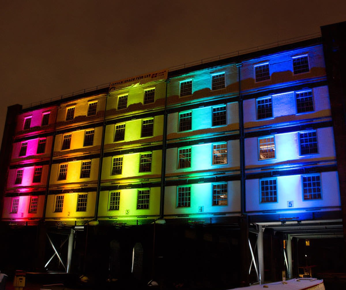 Five-story factory building with each bay lit a rainbow color, windows glowing pink to blue at night