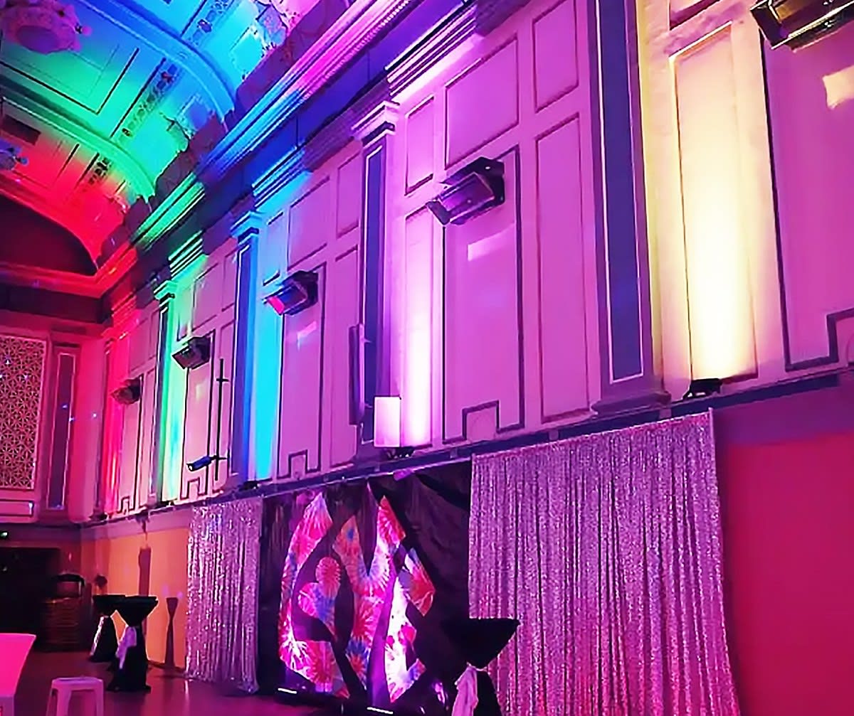 Ballroom walls and ceiling uplit in rainbow colors above sequin drapes and a tie-dye stage backdrop