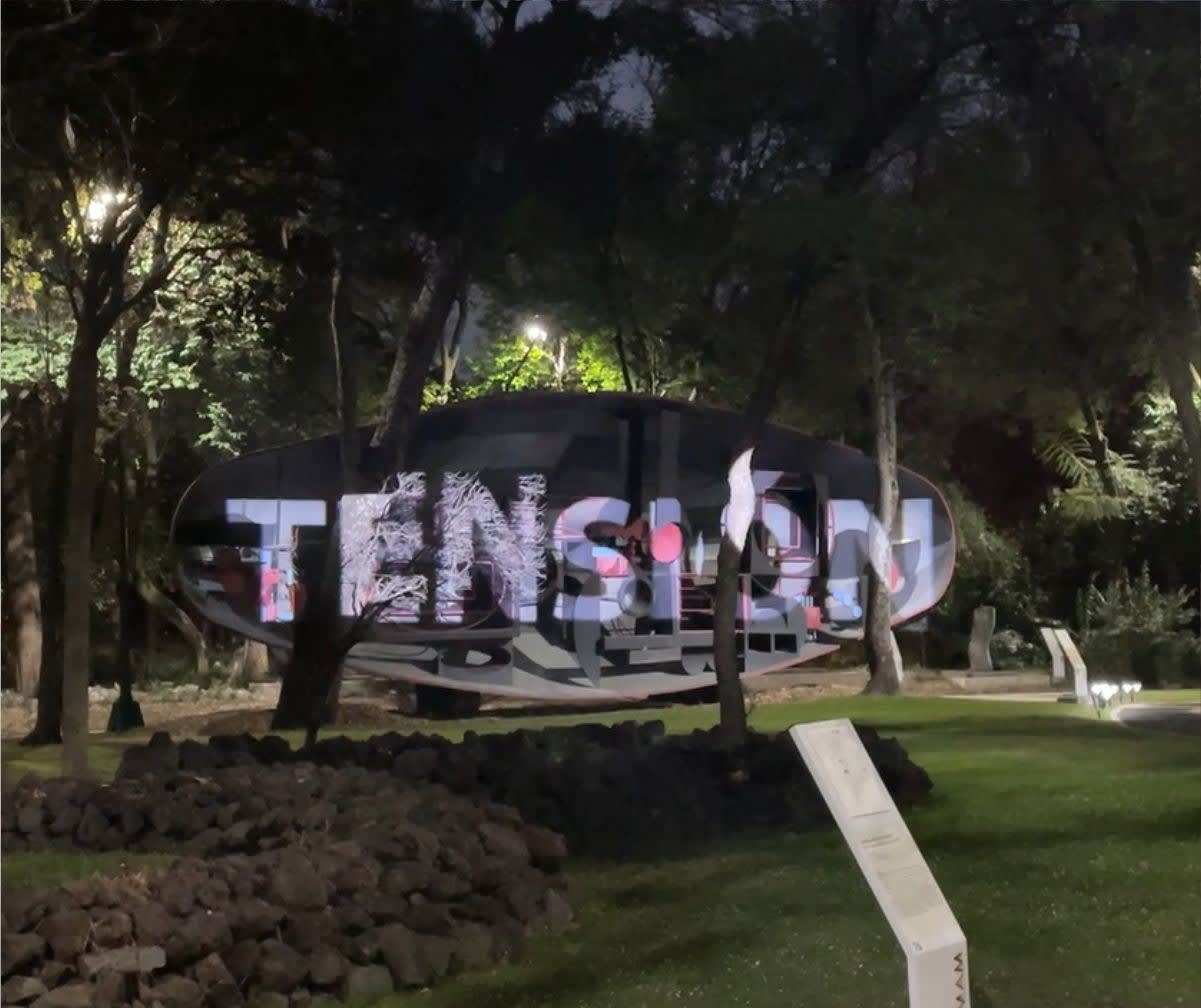 TENSION in tall letters projected onto a curved outdoor pavilion among trees at night; high wattage gobo projector