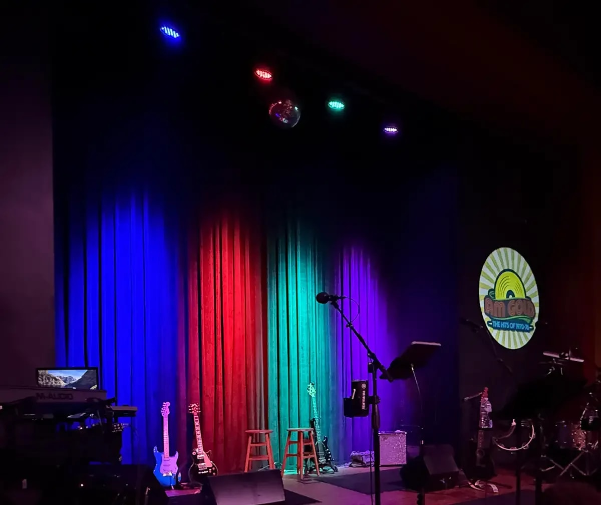 Club stage curtains lit blue, red and green, with guitars waiting and a retro sunburst band logo projected on the wall