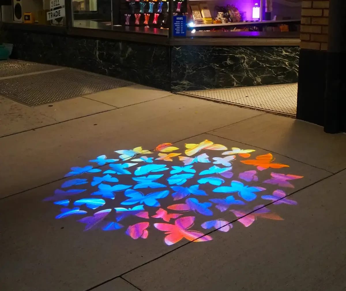 Swarm of blue, pink, and orange butterflies projected on the pavement outside a storefront at night
