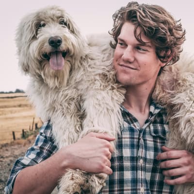 A man with curly hair is embracing a large, shaggy dog in a rural, grassy field with a fence in the background.