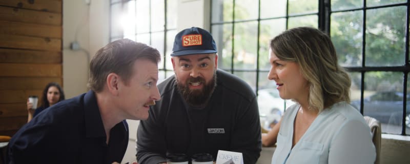 a man holding two coffee cups leans into a table between two people in conversation with each other. he looks into the camera with a pained grimace