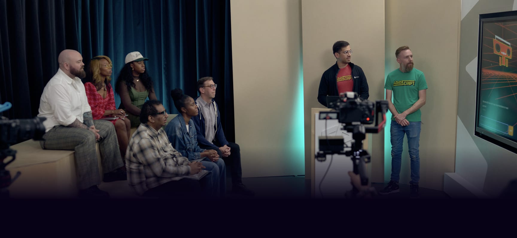A group of developers sitting on bleachers in the Web Dev Challenge demo set, watching two developers present their project