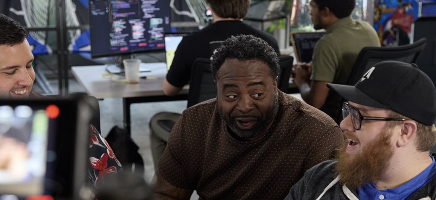 Jason peers over the desk at the code on the screen of Ryan's laptop. Austin is leaning over to see the screen as well