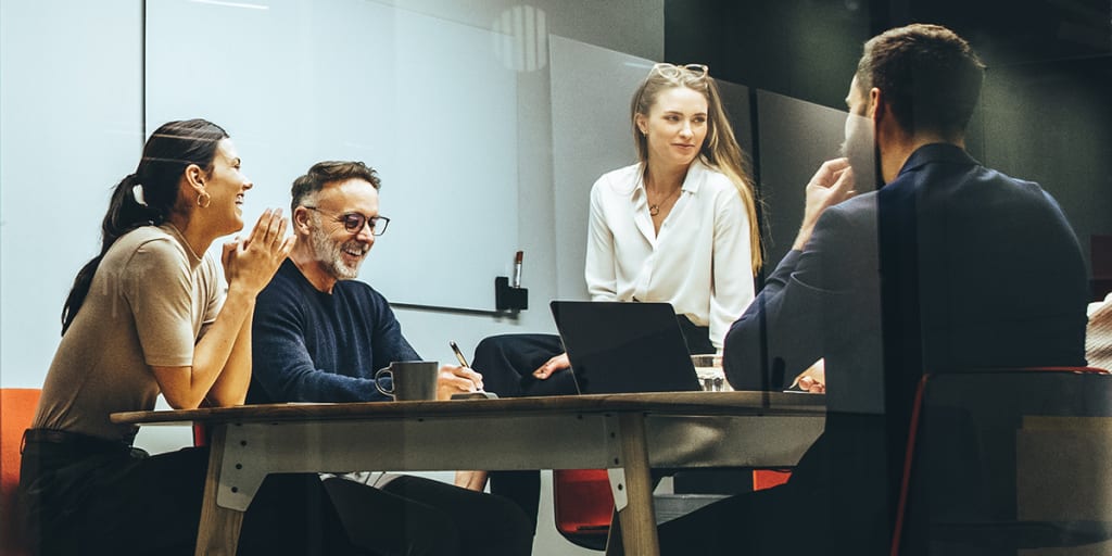 Businesswomen and businessmen sit around conference room table with laptop, having a discussion.