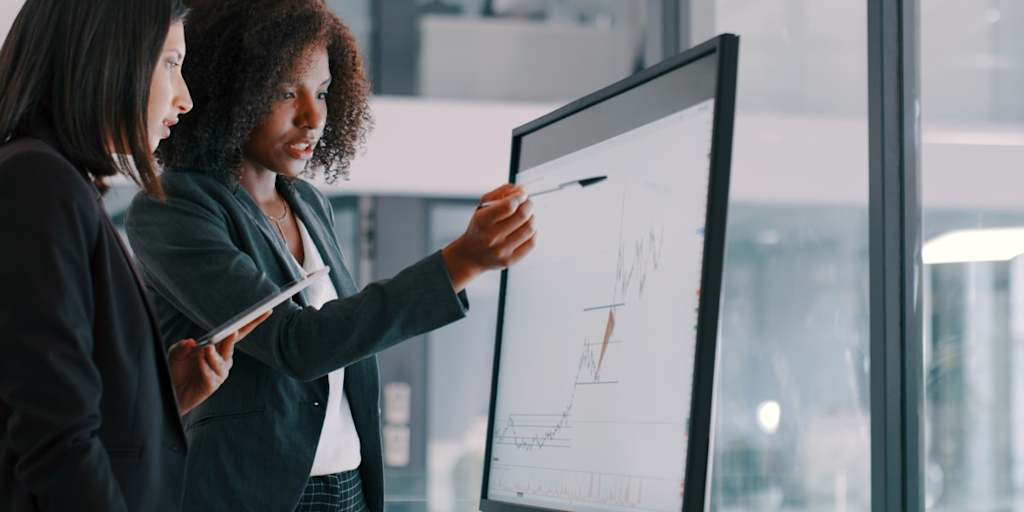 Two professional women review data on a large screen and a mobile device in an office setting