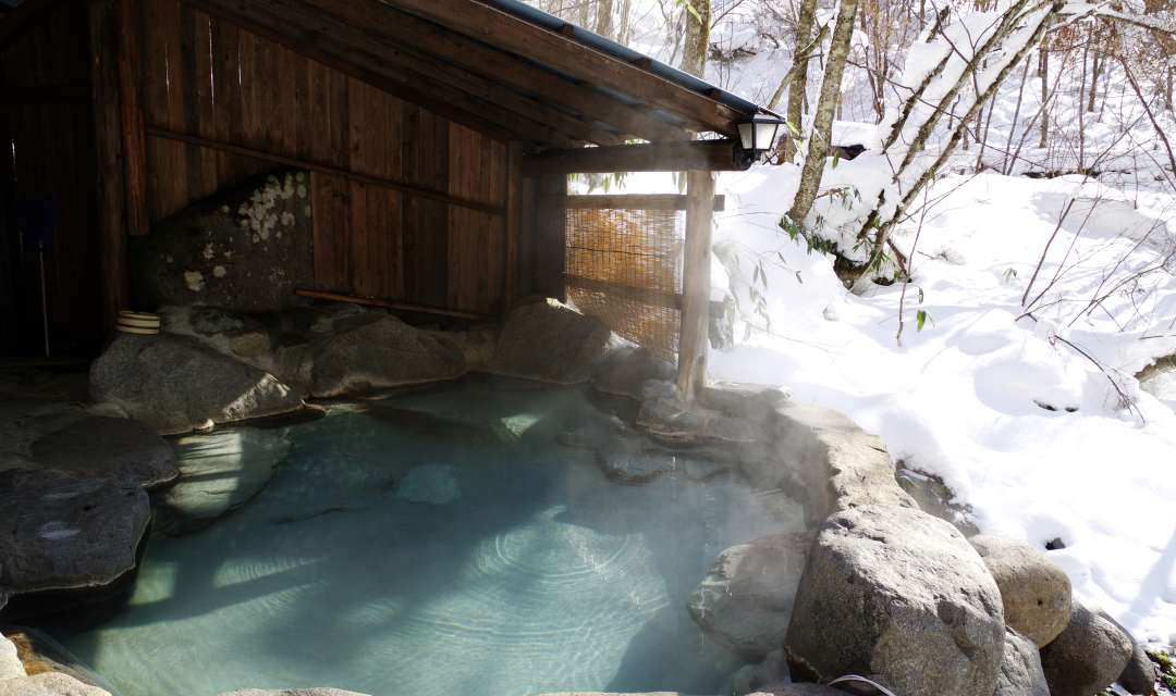 a blue outdoor onsen surorunded by rocks and snow outside a wooden structure